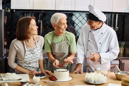 freudige junge Frau in Schürze lächelt Koch und ihren reifen Freund an, der Kuchen mit Beeren dekoriert