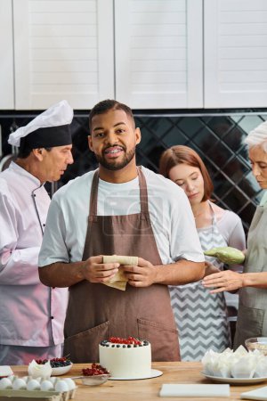 focus on jolly african american man with cake smiling at camera with his blurred friends on backdrop