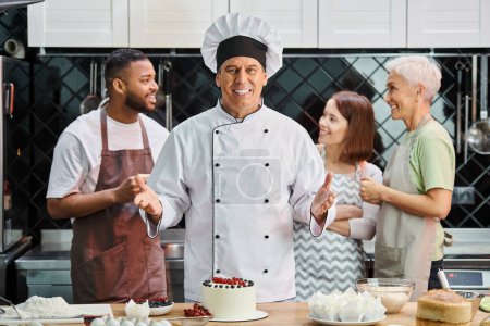 focus on jolly mature chef posing with cake and smiling at camera, his blurred students on backdrop