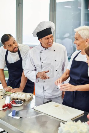 Téléchargez les photos : Joyeux chef cuisinier mature souriant à ses chefs interracial pendant qu'ils cuisinent ensemble, confiserie - en image libre de droit