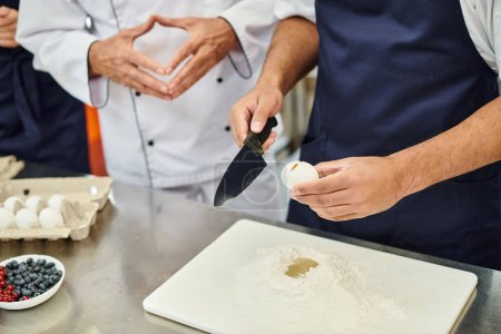 cropped view of african american chef in blue apron breaking egg with knife next to his colleagues