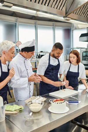 Téléchargez les photos : Joyeux femmes et chef cuisinier en chapeau blanc regarder leur collègue afro-américain casser oeuf - en image libre de droit