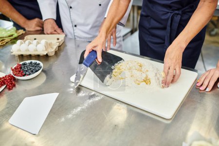 cropped view of mature female chef working with dough next to her colleagues, confectionary