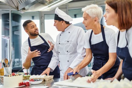 joyous african american chef in apron talking to his colleagues while they working with dough