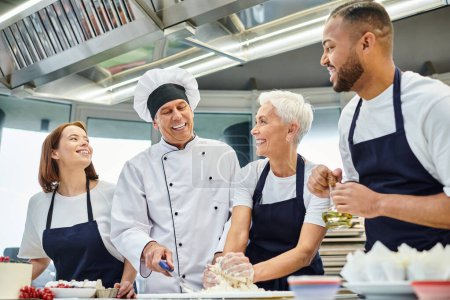Téléchargez les photos : Joyeux chef cuisinier mature en chapeau blanc aidant ses chefs multiculturels avec de la pâte, confiserie - en image libre de droit