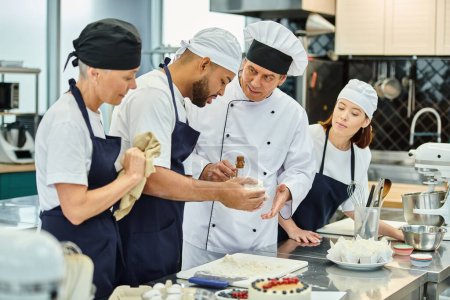Téléchargez les photos : Beau chef cuisinier mature aidant ses chefs multiculturels dans les toques avec de la pâte, confiserie - en image libre de droit