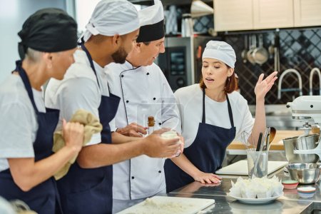 lustige Chefköchin mit weißem Hut lächelt seine junge Köchin an und stellt neben ihren Kollegen Fragen