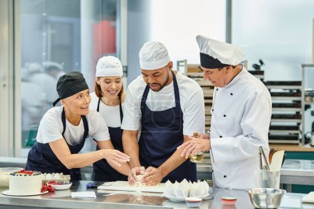 Téléchargez les photos : Joyeux chefs multiculturels dans des tabliers bleus travaillant ensemble avec la pâte à côté du chef cuisinier - en image libre de droit