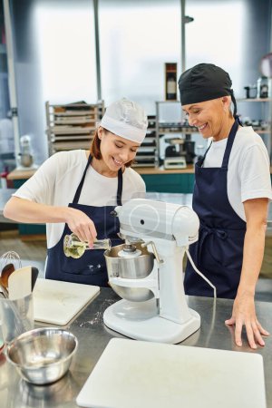 cheerful female chefs in aprons and toques smiling happily and adding oil into mixer, confectionary
