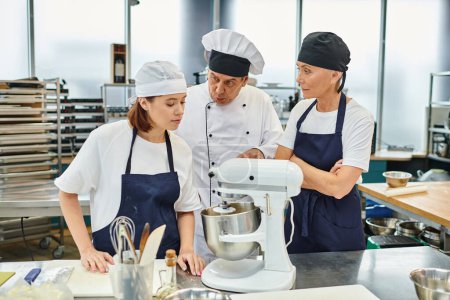 Téléchargez les photos : Beau chef cuisinier mature expliquant comment travailler avec mélangeur à ses femmes chefs, confiserie - en image libre de droit