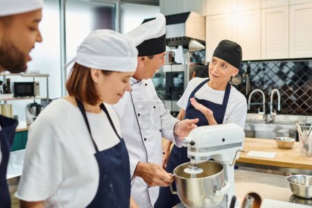 chefs joyeux multiraciaux dans des tabliers bleus et des toques parler avec leur chef cuisinier à côté du mélangeur