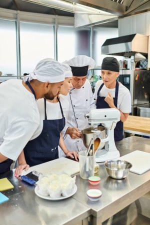 Téléchargez les photos : Beaux chefs multiraciaux dans des tabliers regarder leur chef cuisinier montrant comment travailler avec mélangeur - en image libre de droit