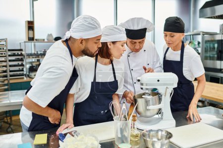 Téléchargez les photos : Chefs multiculturels dans des tabliers bleus et des toques regarder leur chef cuisinier travaillant avec mélangeur - en image libre de droit