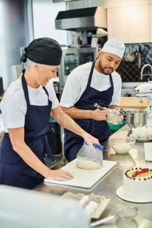 focus on young african american chef in toque whisking next to his blurred jolly mature colleague