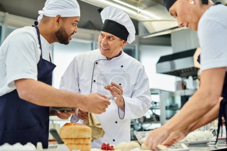 Photo for Cheerful mature chief cook in white hat explaining information joyfully to his multiracial chefs - Royalty Free Image