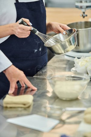 Téléchargez les photos : Vue recadrée du jeune chef féminin dans tablier fouettant des ingrédients pour la pâte à côté du chef cuisinier - en image libre de droit