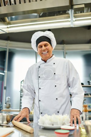 joyous mature chief cook in white hat smiling at camera while baking on kitchen, confectionery