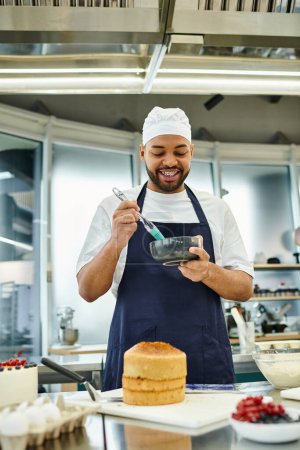 Foto de Alegre joven guapo afroamericano chef en toque usando cepillo de silicona y sonriendo felizmente - Imagen libre de derechos