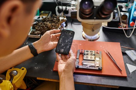 abgeschnittene Ansicht eines erfahrenen Technikers, der ein Handy mit kaputtem Bildschirm in der Nähe von Reparaturgeräten hält