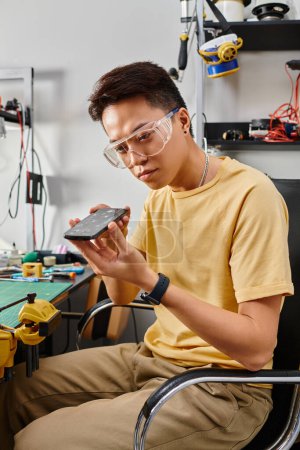 Foto de Reparador asiático en gafas mirando teléfono inteligente durante el mantenimiento en el taller, pequeña empresa - Imagen libre de derechos