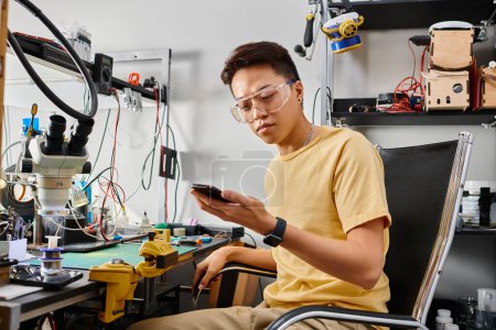 Foto de Experimentado asiático trabajador de taller de reparación mirando el teléfono inteligente durante el diagnóstico técnico - Imagen libre de derechos
