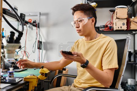 asiatischer Techniker mit Brille hält kaputtes Handy während der Arbeit in privater Werkstatt