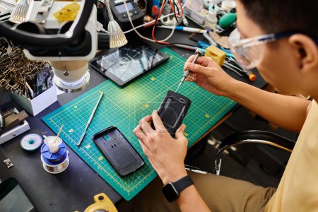 Techniker in Schutzbrille mit Schraubenzieher repariert Handy am Arbeitsplatz in privater Werkstatt