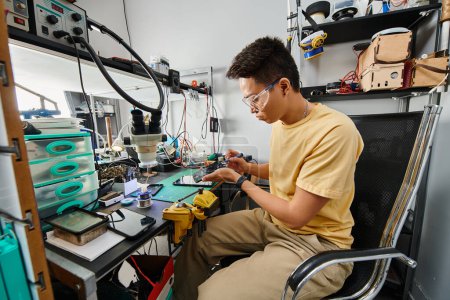 professional asian technician in goggles maintaining electronic devices at workplace in repair shop