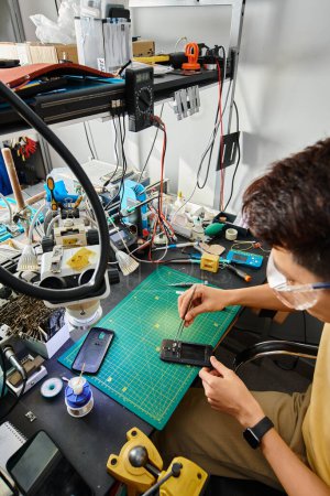 Foto de Experto asiático técnico en gafas de trabajo con roto smartphone y pinzas en su propio taller de reparación - Imagen libre de derechos