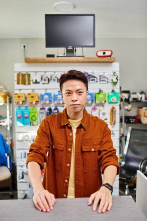 selbstbewusster asiatischer Mann blickt in privatem Elektronikgeschäft mit Elektronik-Sortiment in die Kamera