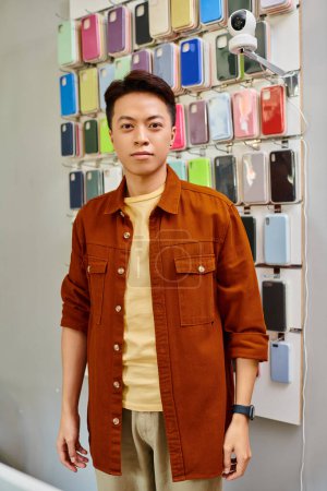 successful asian entrepreneur looking at camera near assortment of phone cases in private shop