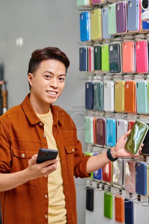 joyous asian man with mobile phone looking at camera near assortment of phone cases in private shop