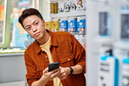 asiático empresario celebración de teléfono móvil y mirando hacia otro lado en propia tienda de electrónica, pequeña empresa