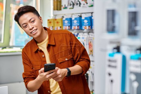 froh asiatische Ladenbesitzer hält Handy und schaut weg in Elektronik-Geschäft, kleines Geschäft