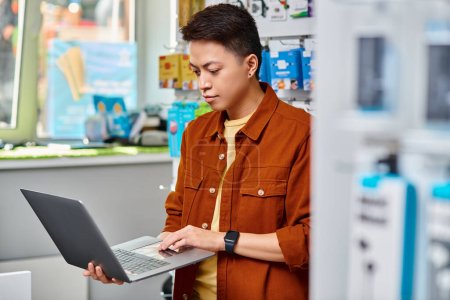 konzentrierter asiatischer Mann mit Laptop, während er in einem privaten Elektronikgeschäft steht, Kleinunternehmen