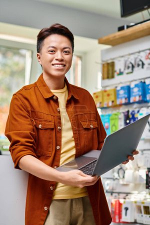 joyful asian store owner with modern laptop looking at camera in electronics shop, small business