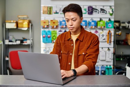 serious asian man using laptop on counter in private electronics store, small business concept