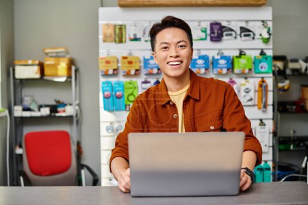 joyful asian electronics store owner smiling at camera near laptop in own shop, small business