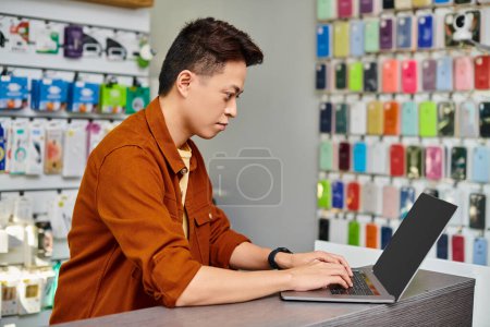 Photo for Concentrated asian man using laptop on counter in private electronics shop, small business concept - Royalty Free Image