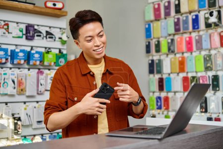 Foto de Sonriente asiático empresario mostrando smartphone durante videollamada en portátil en tienda privada - Imagen libre de derechos
