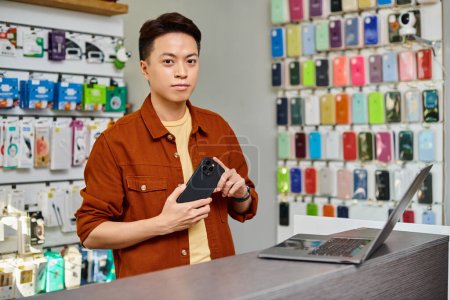 vendedor asiático seguro de sí mismo con el teléfono móvil mirando a la cámara cerca de portátil en el mostrador en la tienda privada