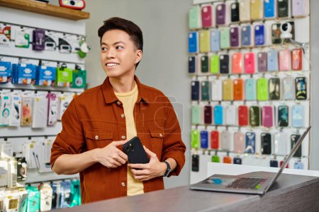 happy asian businessman with mobile phone looking away near laptop in private electronics shop