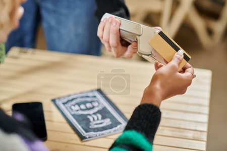 young customer holding credit card near card reader, cropped hand on woman paying in vegan cafe