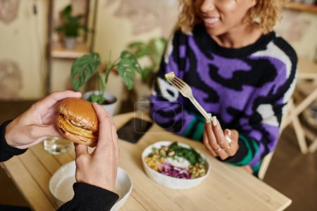 vista recortada de pareja joven y diversa disfrutando de comida vegana en la cafetería, hamburguesa con tofu y ensalada