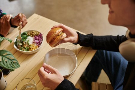 hombre joven recortado disfrutando de comida vegana mientras sostiene hamburguesa con tofu cerca de la mujer borrosa comer ensalada