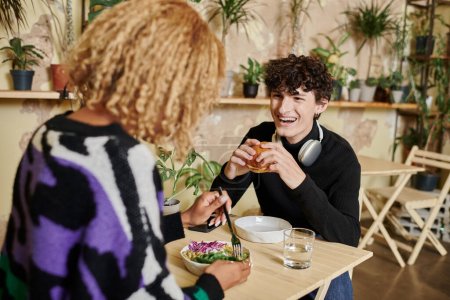 happy curly man eating tofu burger near african american girlfriend enjoying salad in vegan cafe