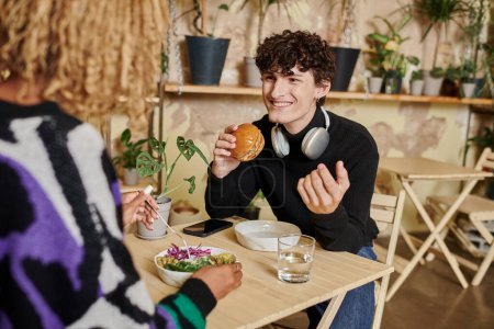 glücklicher junger Mann mit Kopfhörern hält Tofu-Burger in der Hand und schaut eine Afroamerikanerin im Café an