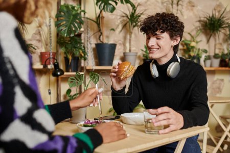 glücklicher junger Mann mit Kopfhörern schaut Tofu-Burger neben glücklicher Afroamerikanerin in veganem Café an