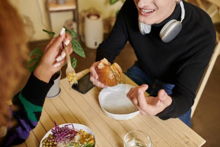 cropped shot of happy man in wireless headphones eating vegan tofu burger near woman in cafe