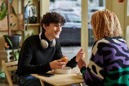 happy curly man with wireless headphones holding tofu burger and chatting with girl in vegan cafe